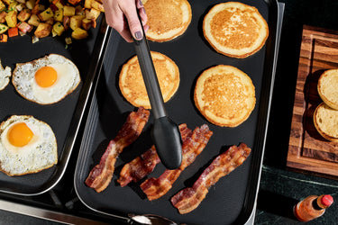 A person cooking eggs, bacon, and pancakes on the HexClad Double Burner Griddle.