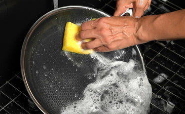 A person scrubbing a HexClad pan under running water with a soapy yellow sponge.